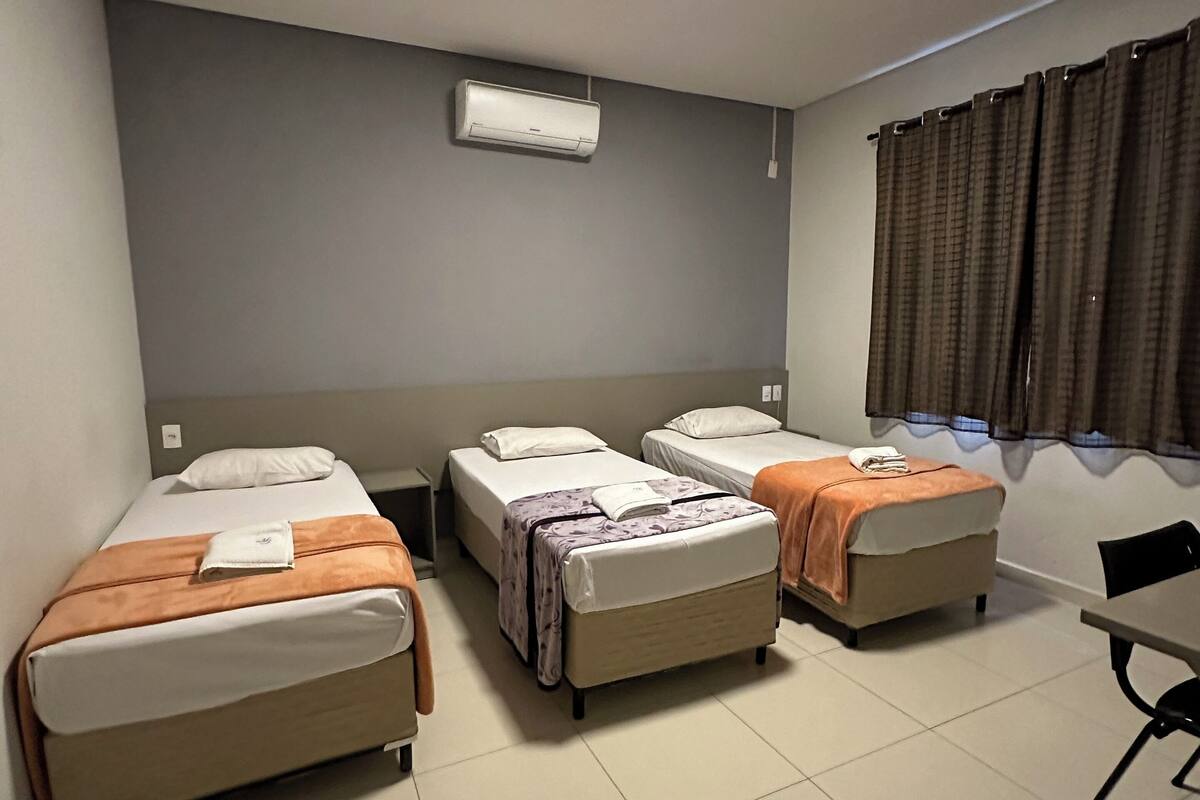Ein Hotelzimmer mit drei Betten, einem Deckenventilator, einer Klimaanlage und einem Vorhangfenster.