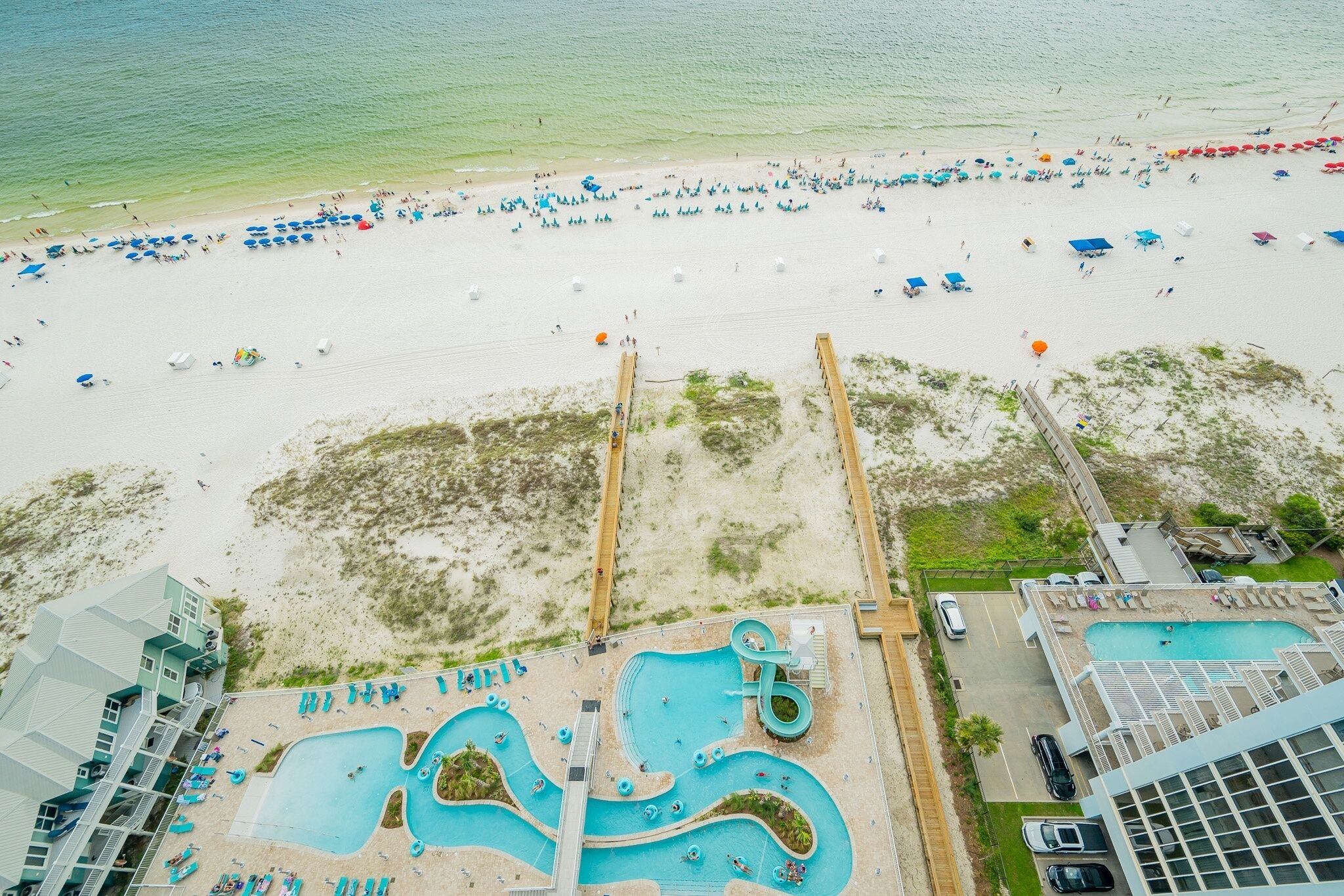 An aerial view of a beach resort with a swimming pool area, beach umbrellas, and a wooden boardwalk.