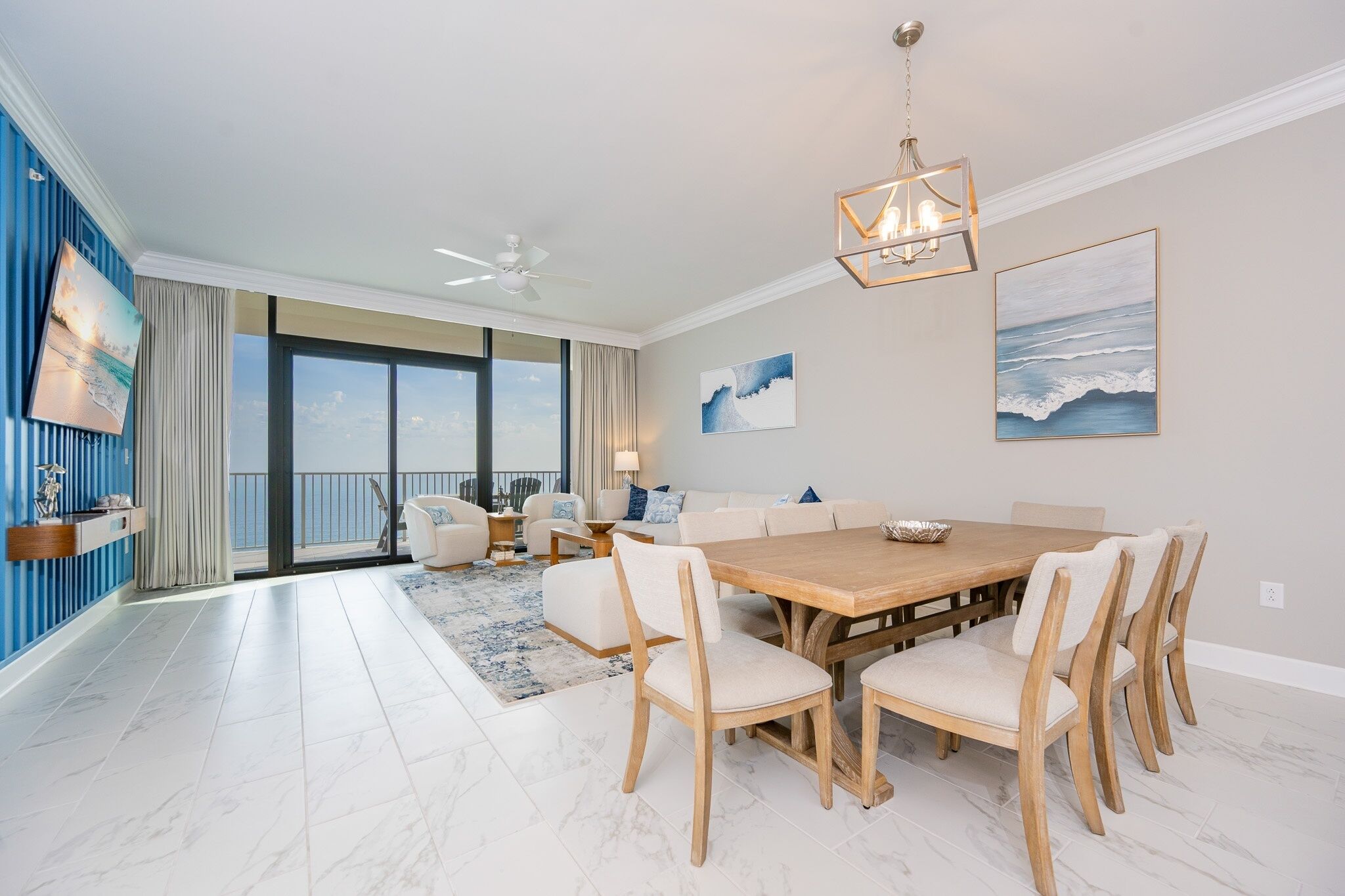 A spacious living room with a large wooden dining table, white upholstered chairs, a ceiling fan, and a view to the outside.