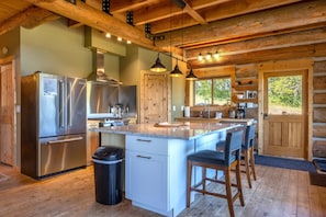 A log cabin kitchen with a stainless steel refrigerator, wooden cabinets, a large island with bar stools, pendant lighting, and a wooden door. - Adams Rest 3 Bedroom Cabin (The Dalles)