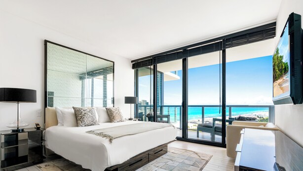 Un dormitorio moderno con una cama grande, un televisor y un balcón con vistas a la playa y al océano.