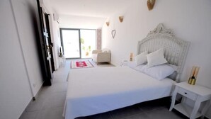 A bedroom with a large bed, a bedside table, a nightstand, a sliding glass door leading to a balcony, and a small seating area with a chair and table.