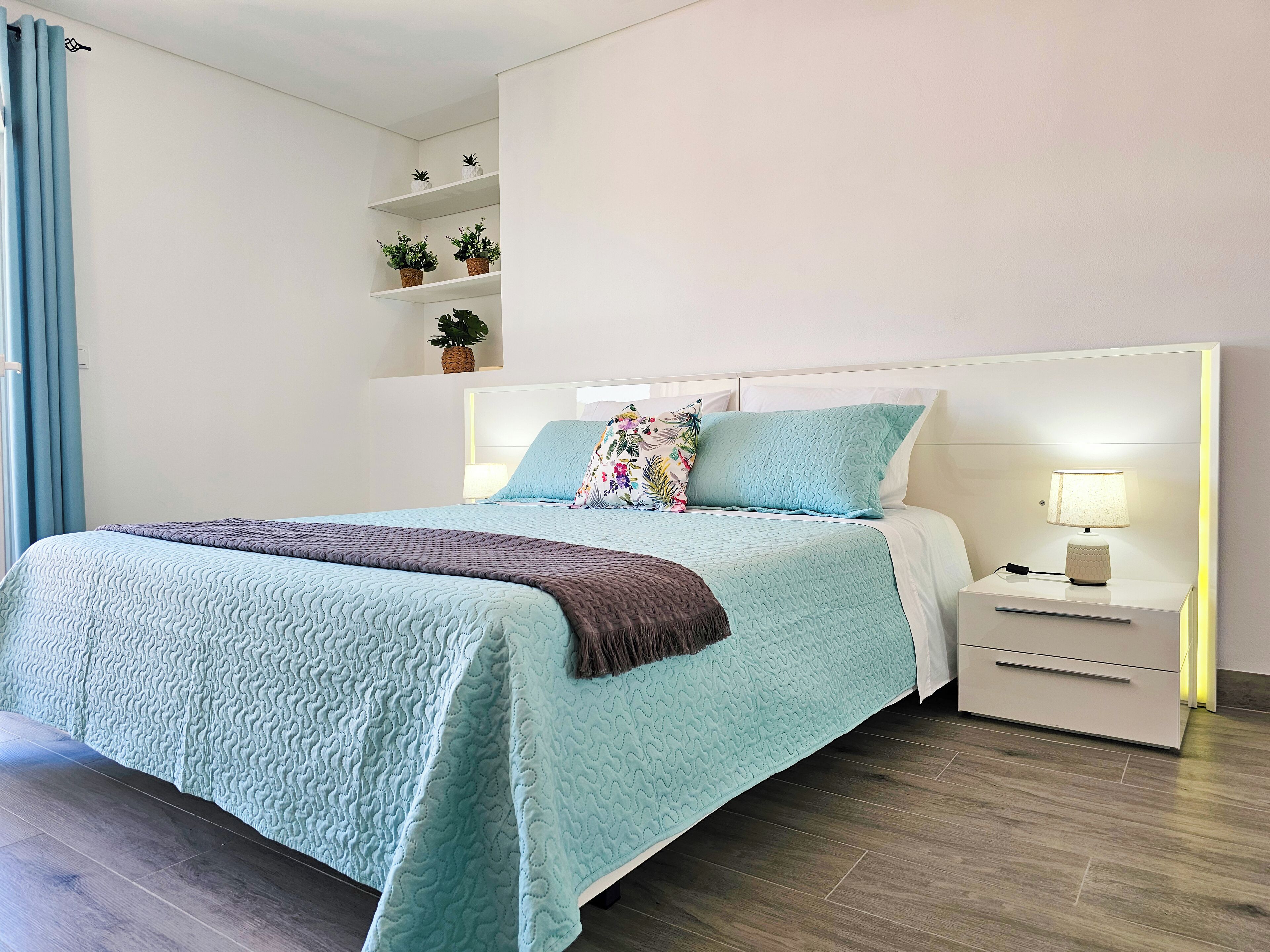 Un lit bien fait, recouvert d’une courtepointe turquoise, d’un plaid marron et agrémenté d’un coussin à motifs floraux. Une table de chevet avec une lampe et une petite plante se trouve à côté du lit. La chambre est dotée d’une tête de lit blanche et d’une étagère où sont disposées des plantes en pot.