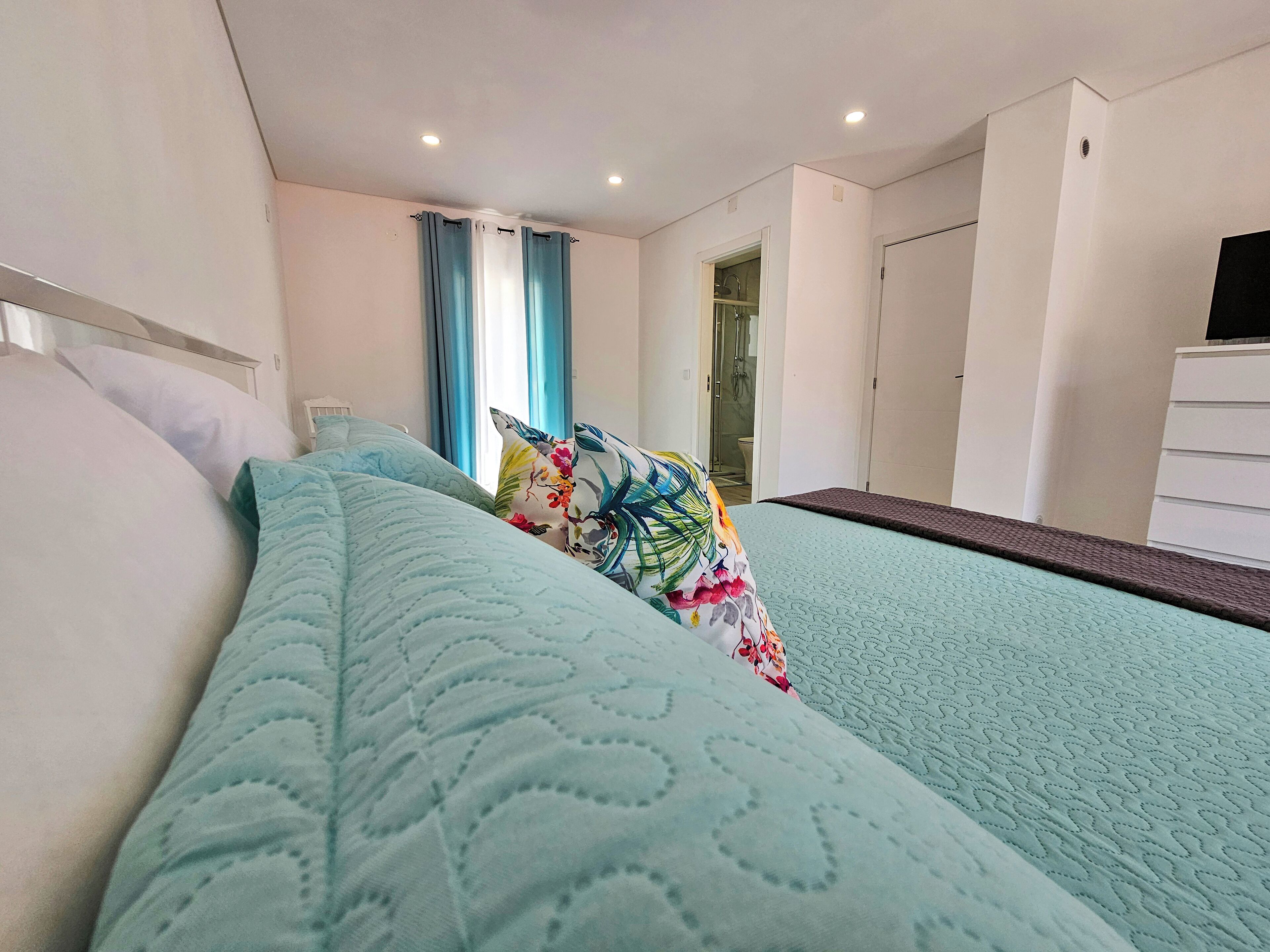 Un letto rifatto con cura, con una coperta e dei cuscini a motivi, un comò bianco e un bagno con doccia visibile attraverso una porta aperta.