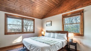 Un dormitorio con techo de madera, dos ventanas amplias, una cama con ropa blanca, una mesita de noche con lámpara y una alfombra estampada.