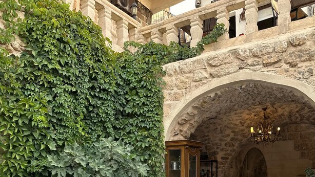 A courtyard with a stone wall covered in greenery, a wooden cabinet, and a seating area with patterned cushions. - ACABEY KONAĞI (Mardin)