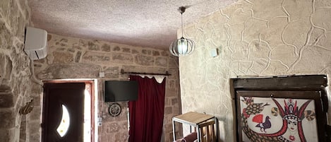 Una stanza rustica con parete in pietra, un letto con coperta rossa, un piccolo armadietto bianco, una ventola a soffitto e una TV a muro.
