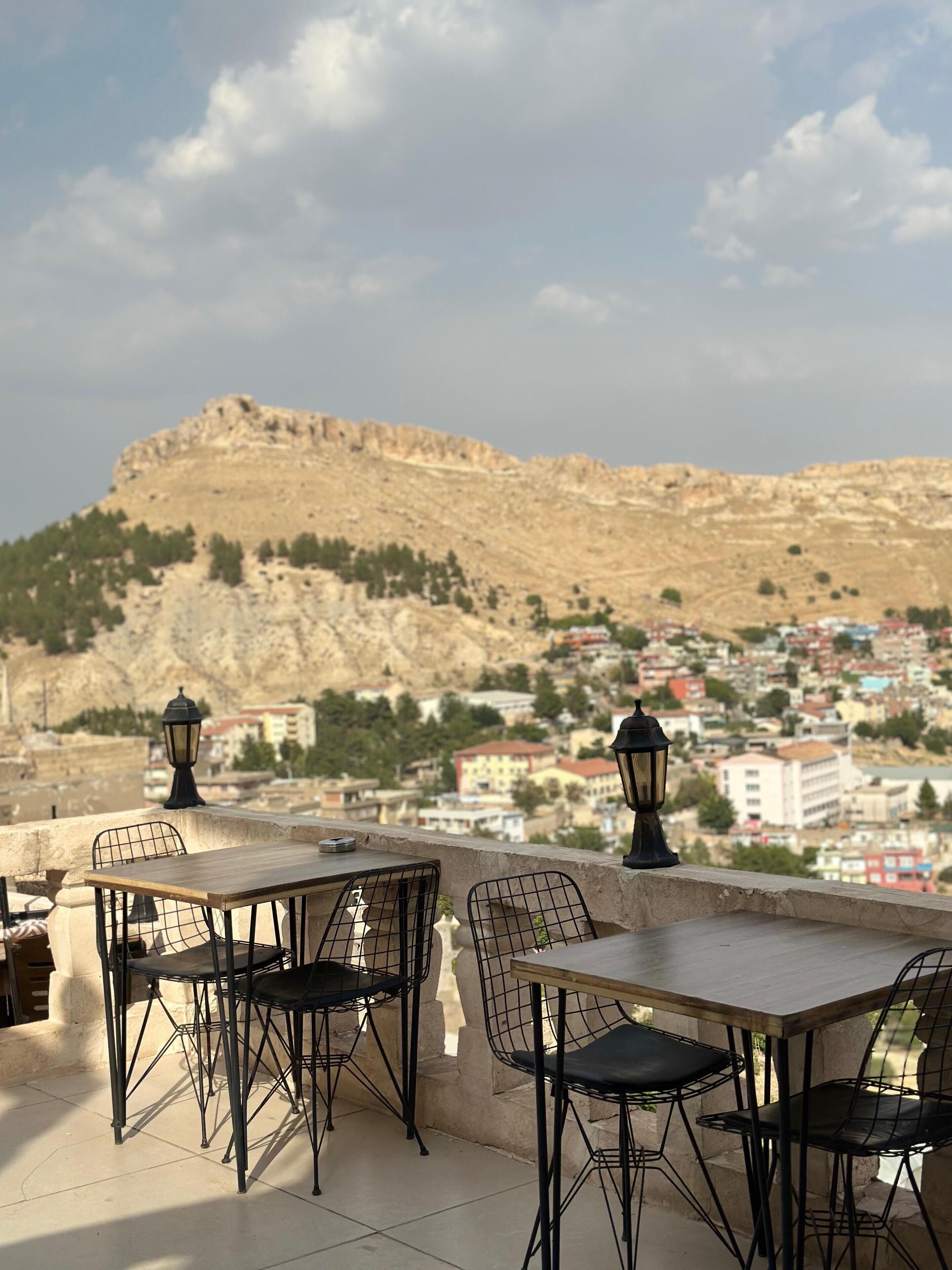 An outdoor terrace with tables and chairs, overlooking a town and a hill.