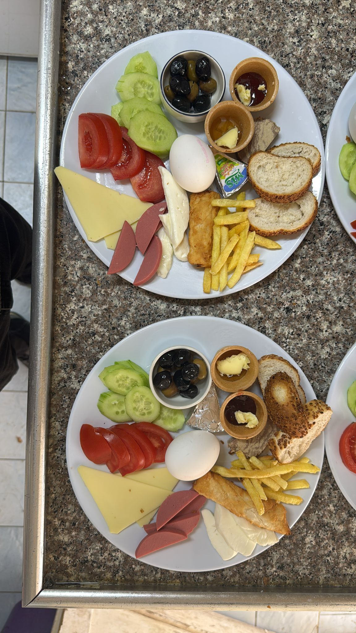 Prato de pequeno-almoço com vários itens, incluindo ovos, queijo, fiambre, tomates, pepinos, azeitonas, pão e batatas fritas.