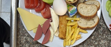 A breakfast plate with various items including eggs, cheese, ham, tomatoes, cucumbers, olives, bread, and French fries.