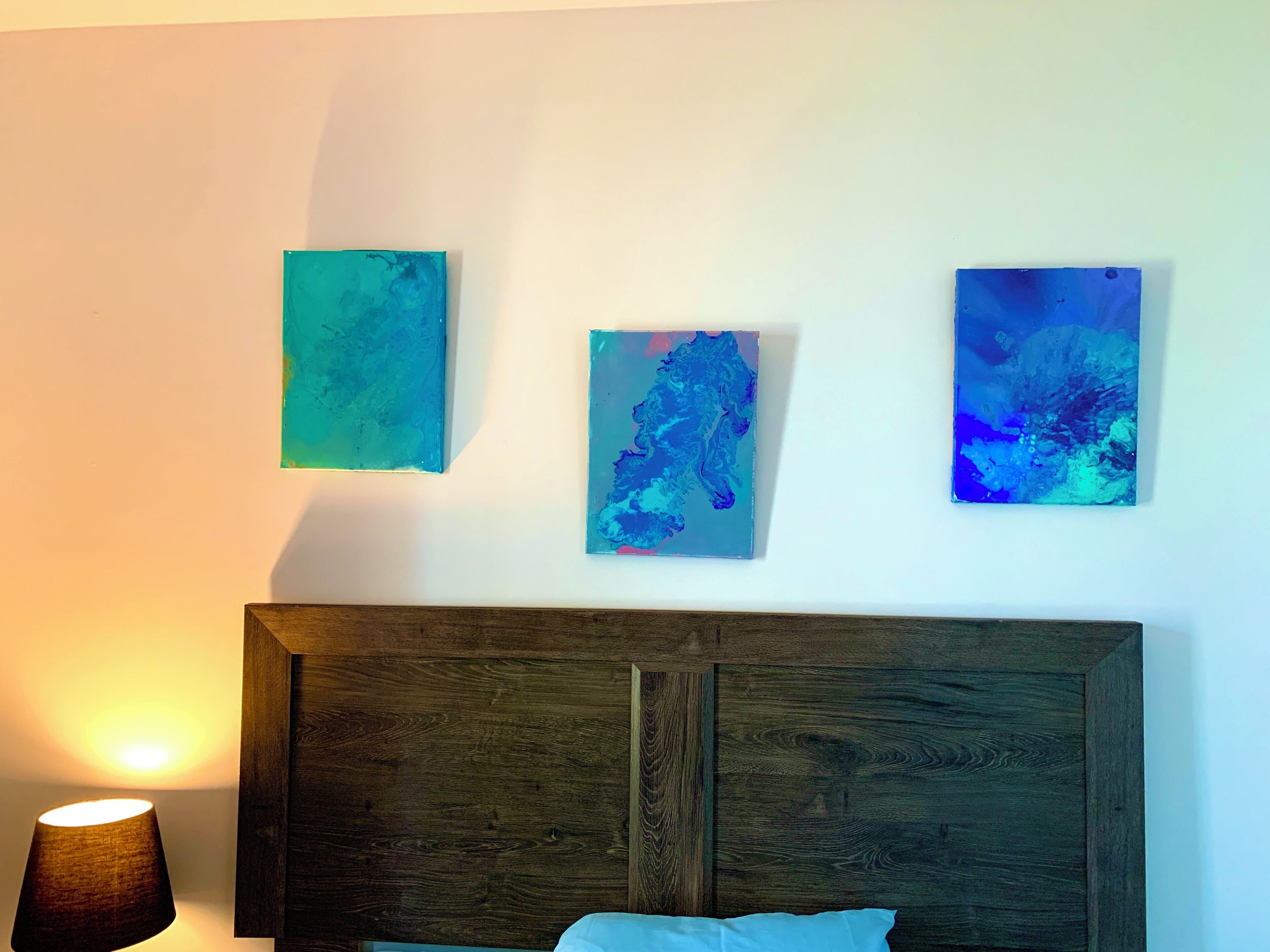 Quarto com cabeceira de madeira, uma luminária e três quadros abstratos na parede.