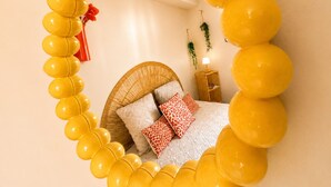 Una habitación con cama, una silla de mimbre, una mesita y un estante en la pared.