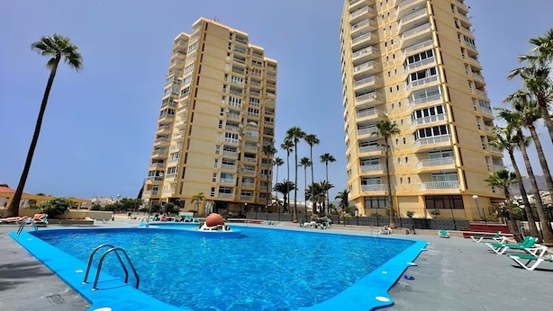 A swimming pool area with a blue pool, surrounded by palm trees and two tall residential buildings. - Troya Beach Apartments (Playa de la Américas)