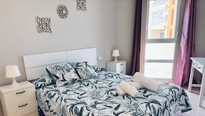 A neatly made bed with a leaf-patterned bedspread, a bedside table with a lamp, and a window with curtains. - Troya Beach Apartments (Playa de la Américas)