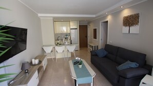 A modern living room with a dark sofa, a TV mounted on the wall, and a dining area with white chairs and a small table. - Troya Beach Apartments (Playa de la Américas)