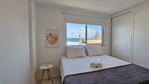 A bedroom with a bed, a window with a beach view, a small round table, and a wall-mounted artwork.