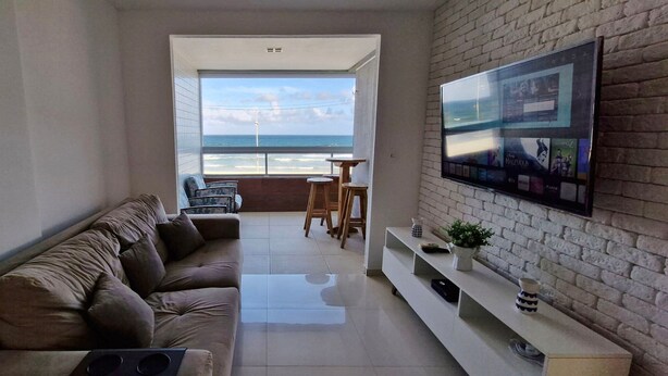 A modern living room with a sofa, a TV mounted on a brick wall, and a view of the ocean through a large window.