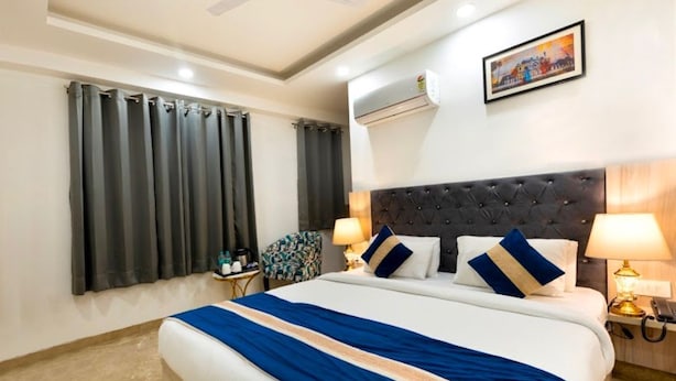 A hotel room with a large bed, two bedside lamps, a headboard, a nightstand, a chair, a window with curtains, an air conditioning unit, and a painting on the wall. - Hotel Evergreen Stay (New Delhi)