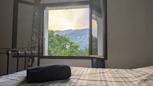 Una cama con una manta escocesa y una almohada negra, una ventana que ofrece vistas a árboles verdes y montañas, y una barandilla de metal.