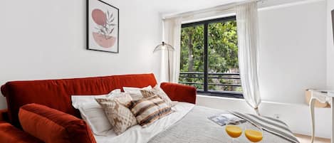 A bedroom with a large bed, a window with curtains, a bedside table, and a tray with breakfast items.