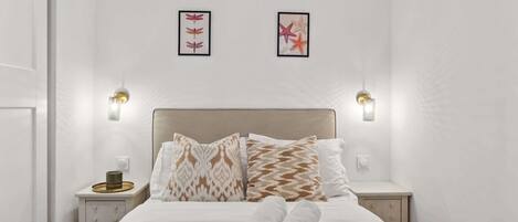 Una cama bien tendida con almohadas estampadas, una toalla doblada y una mesita de noche con una taza y una bandeja.