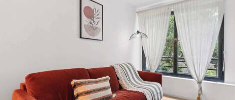 A red velvet sofa with striped and solid cushions, a framed abstract art piece, a floor lamp, and a window with sheer curtains.