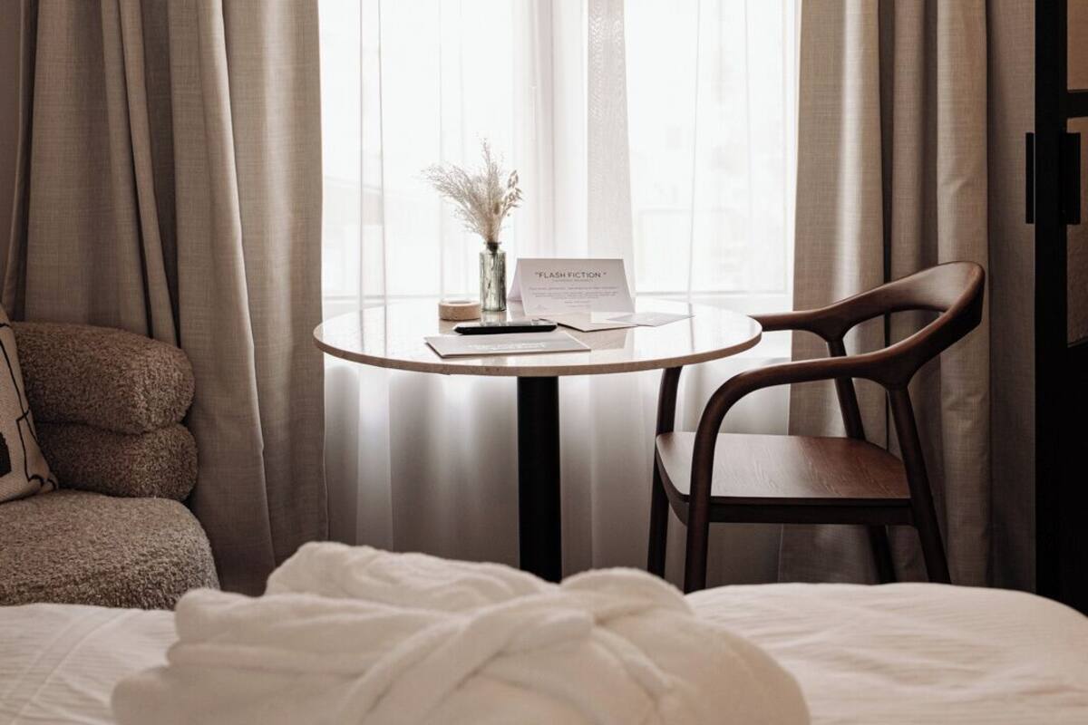 Une chambre d’hôtel avec un lit, une chaise, une table sur laquelle se trouve un livre et un vase de fleurs, ainsi que des rideaux légers.