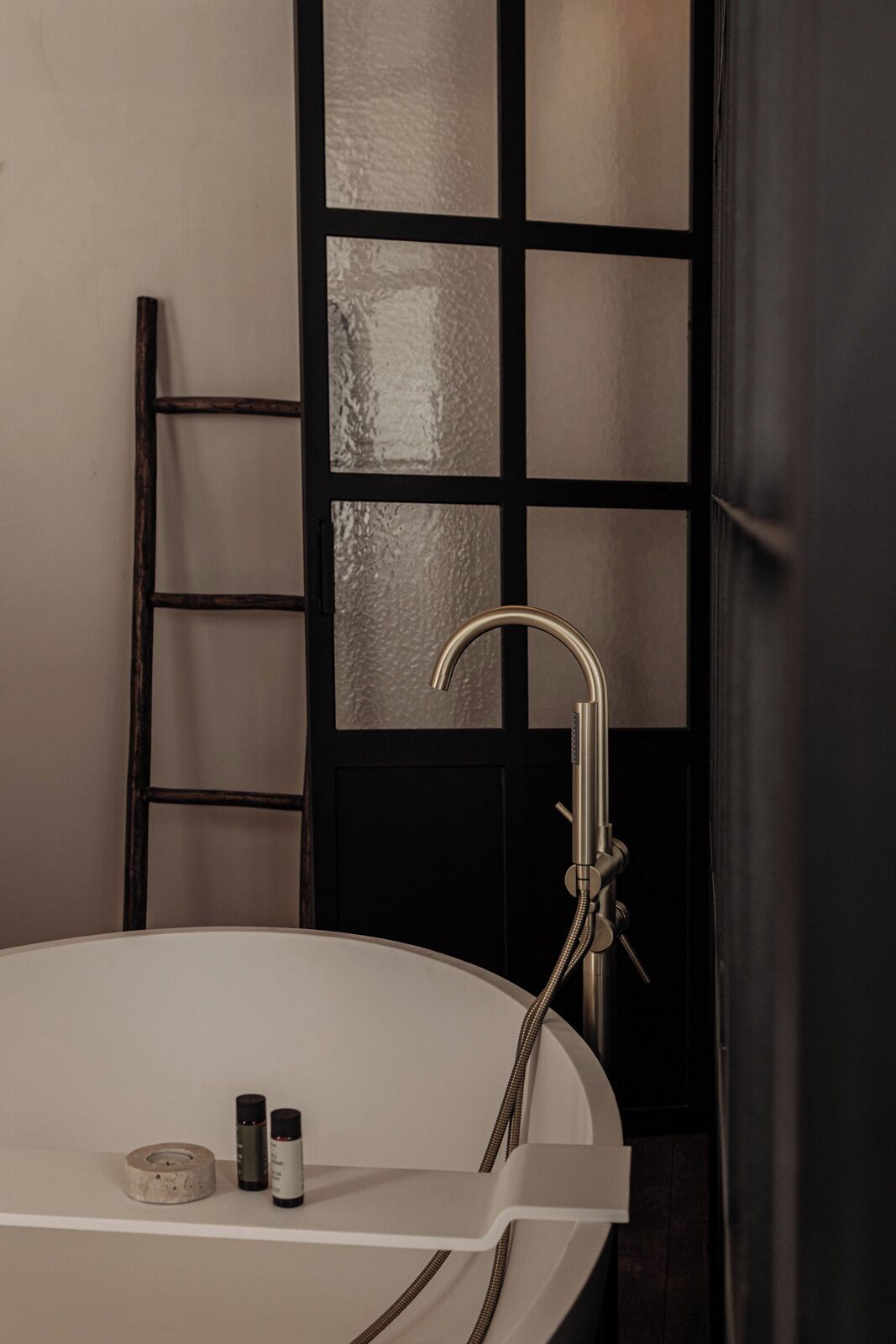 Ein modernes Badezimmer mit einer freistehenden Badewanne, einem Metallarmaturen und einer Holzleiter, die an einer strukturierten Wand lehnt.