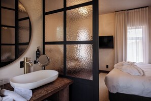 Une chambre d’hôtel dotée d’un grand lavabo ovale, d’un miroir, d’un lit avec du linge de maison blanc et d’un téléviseur fixé au mur.