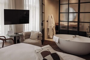 A hotel room with a large bathtub, a television, a cozy armchair, and a desk with a sign.