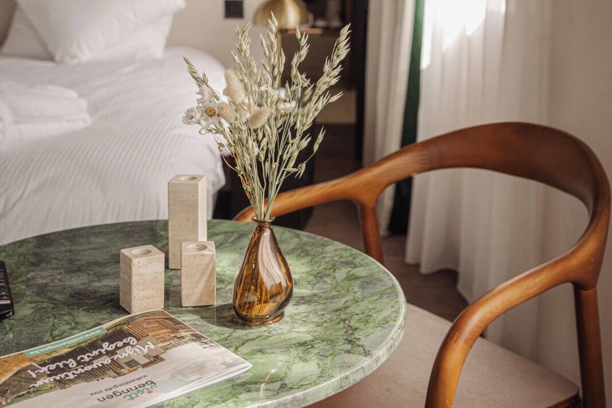 Une chambre d’hôtel avec un lit, une chaise en bois, une table en marbre surmontée d’un vase et un magazine.