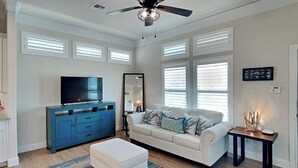 A modern living room with a white sofa, a blue cabinet, a flat-screen TV, and a ceiling fan. - Coastal Zest | Pet-friendly, Private Pool (Port Aransas)