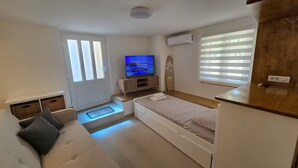 Ein gemütlicher Raum mit einem Bett, einem Fernseher auf einem Tisch, einem Fenster mit Jalousien und einer kleinen Küchenzeile.