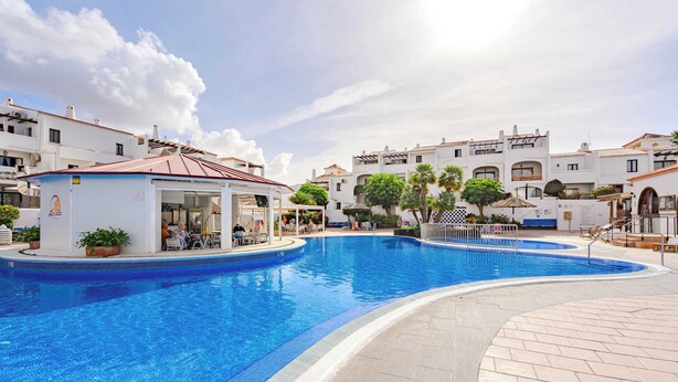 Una piscina con sombrilla, rodeada de edificios con balcones y un cielo despejado.