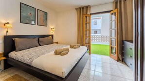 Un dormitorio con cama, cómoda, mesita de noche, lámpara y vistas a un césped verde y un edificio exterior.