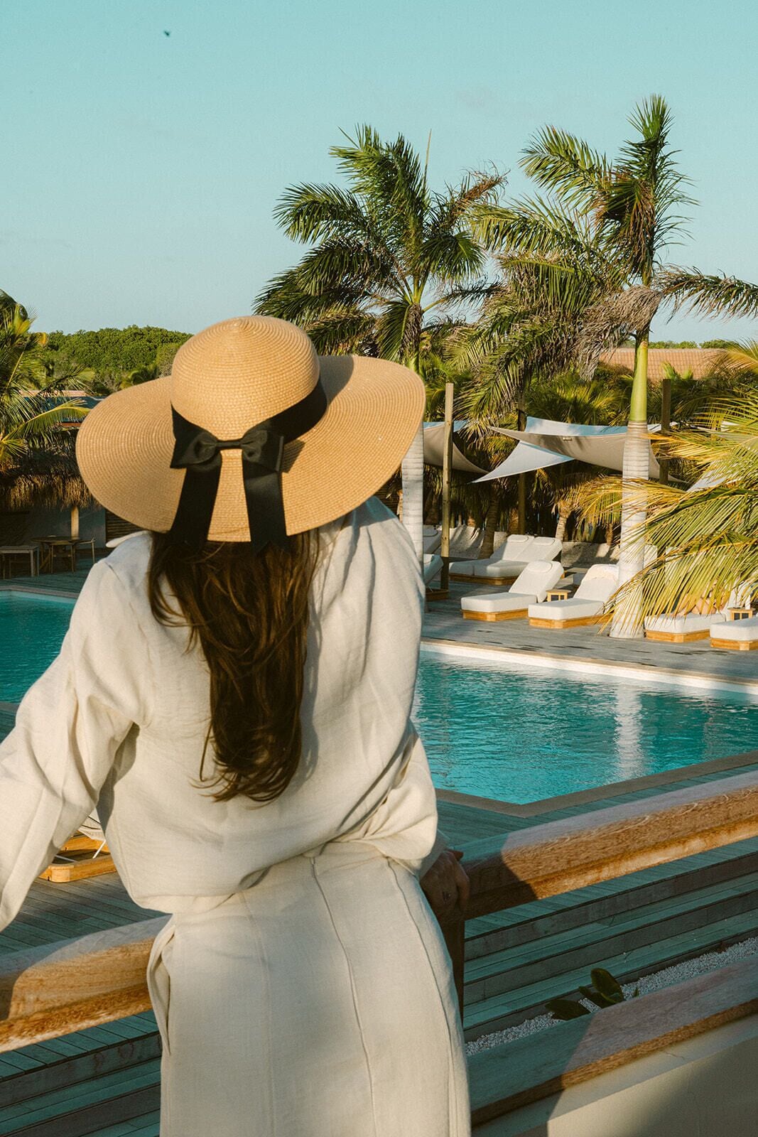 A person with a large sun hat overlooking a pool area with lounge chairs and palm trees.