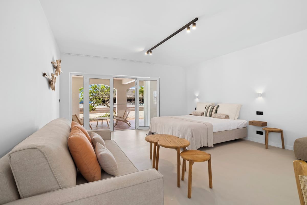 A modern hotel room with a sofa, a bed, a small table, and a view of the beach through large sliding doors.