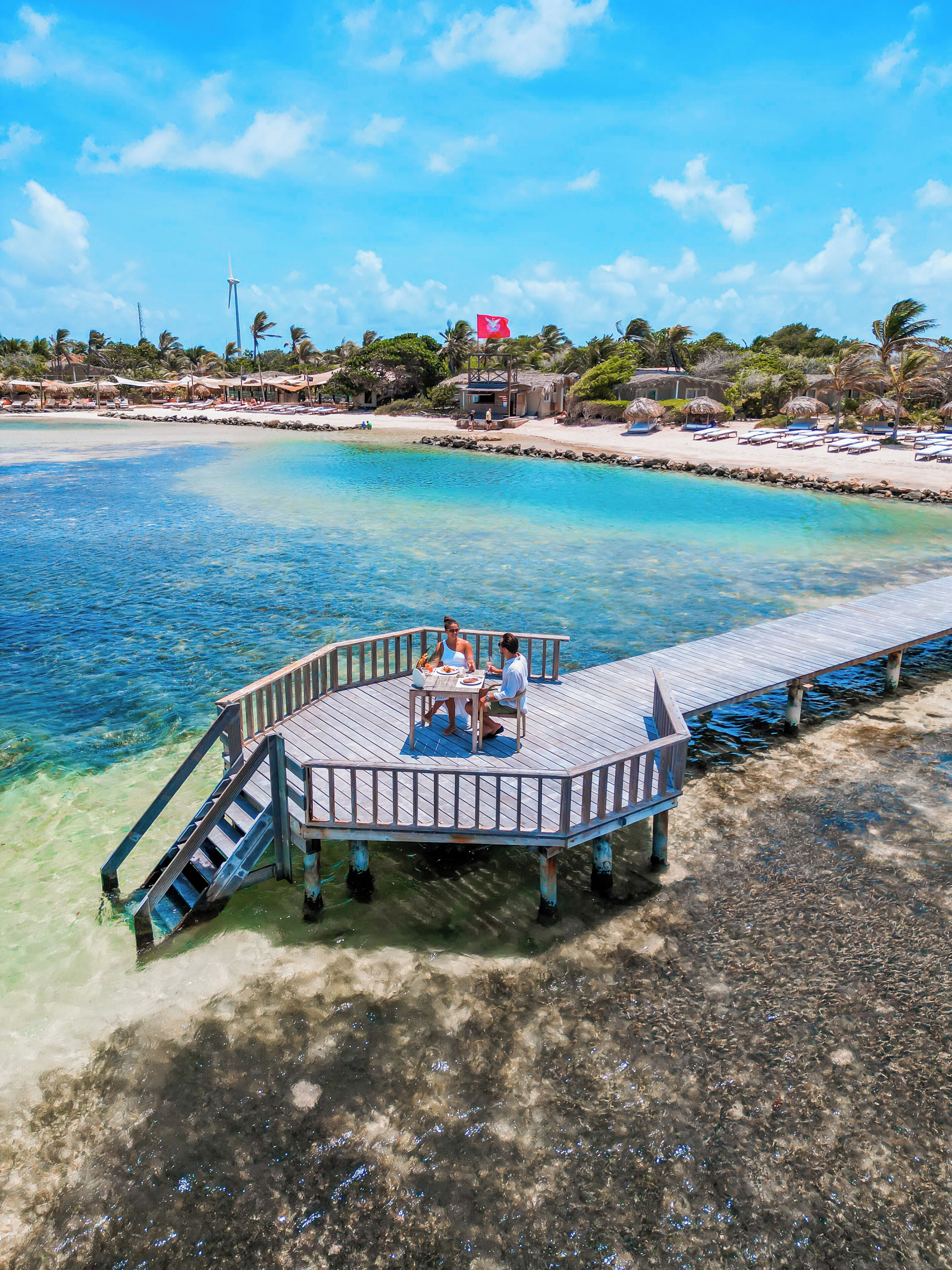 Une terrasse en bois avec une table et des chaises, d’où l’on peut admirer une mer d’un bleu limpide et une plage de sable bordée de palmiers.