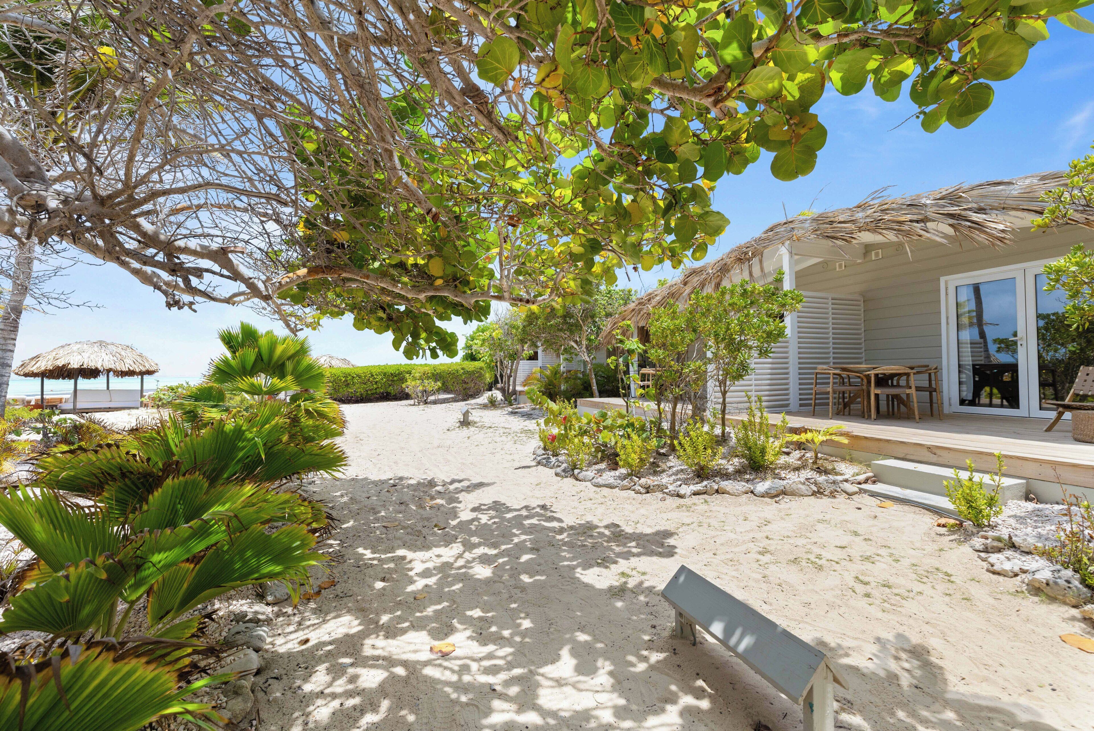 Una propiedad frente al mar con un sendero de arena, una estructura de techo de paja y una terraza de madera con mobiliario exterior.