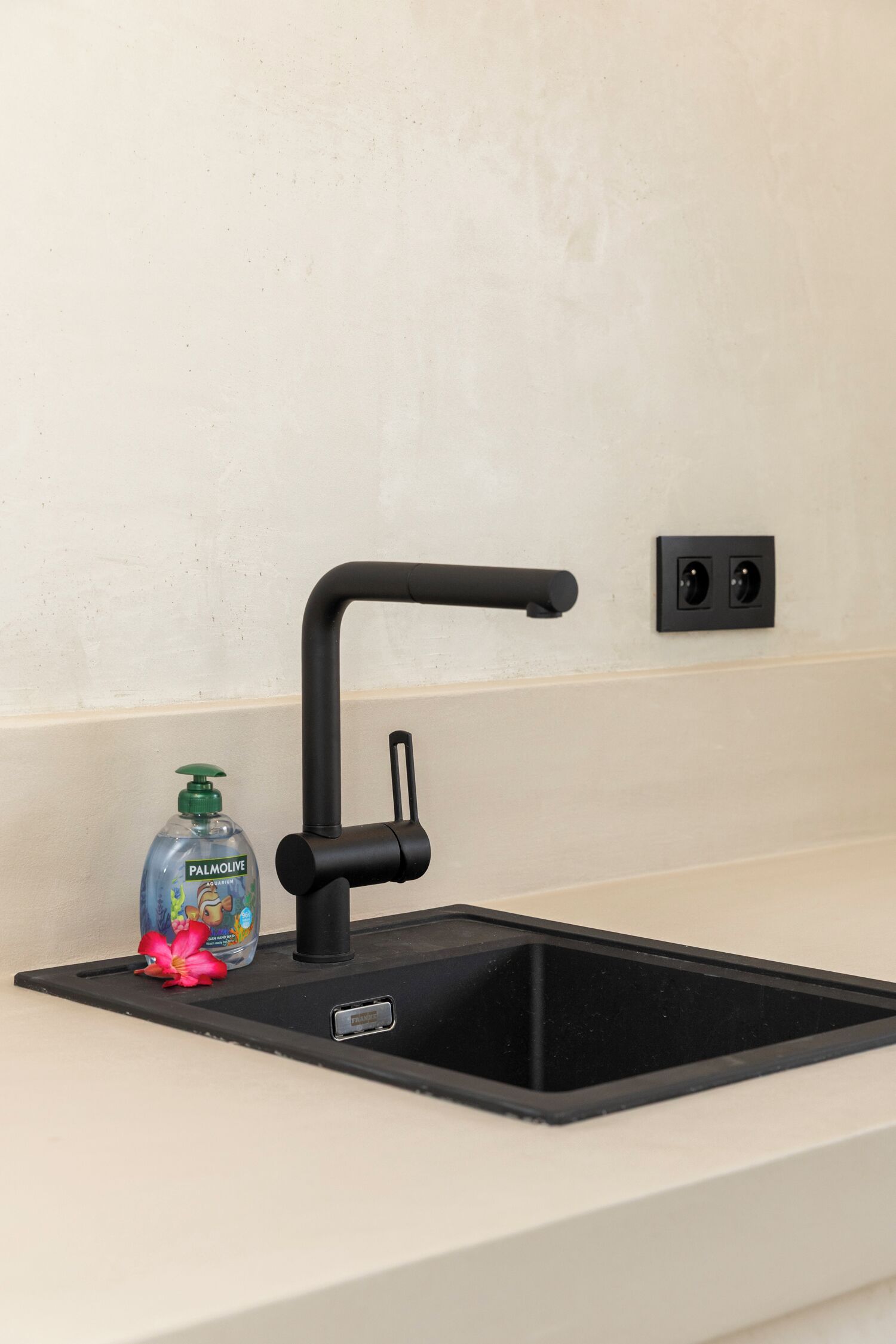 A modern kitchen sink with a black faucet, a soap dispenser, and a power outlet on the wall.