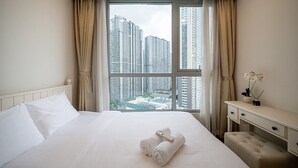 A hotel room with a large window, a bed with white linens, a bedside table with a vase of flowers, and a view of high-rise buildings.