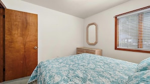 A bedroom with a wooden door, a bed with a floral bedspread, a wicker dresser, a mirror, and a window with blinds.