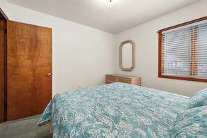 A bedroom with a wooden door, a bed with a floral bedspread, a wicker dresser, a mirror, and a window with blinds.