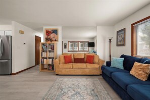 A living room with a beige sofa, a blue sofa, a bookshelf, a rug, and a window with blinds.