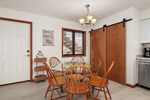 A dining area with a glass table and wooden chairs, a chandelier, a sliding barn door, and a kitchen with a coffee maker.