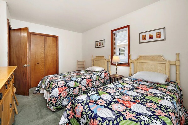 A bedroom with two beds, a wooden dresser, a window with blinds, and framed pictures on the wall.
