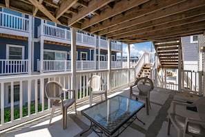 Un espacio exterior cubierto con una mesa y sillas de vidrio, barandillas de madera y vistas a un edificio azul con balcones.