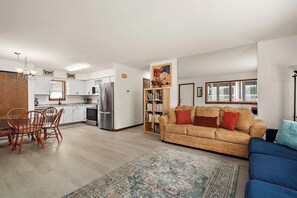 Ein modernes Wohnzimmer mit einer Couch, einem Essbereich mit Stühlen, einer Küche inklusive Geräte und einem Bücherregal.