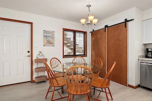 Uma sala de jantar com mesa de vidro e cadeiras de madeira, um lustre, uma porta de celeiro deslizante e uma cozinha com cafeteira.