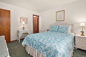Quarto com cama, mesa de cabeceira, cadeira, porta e quadro fixado na parede.